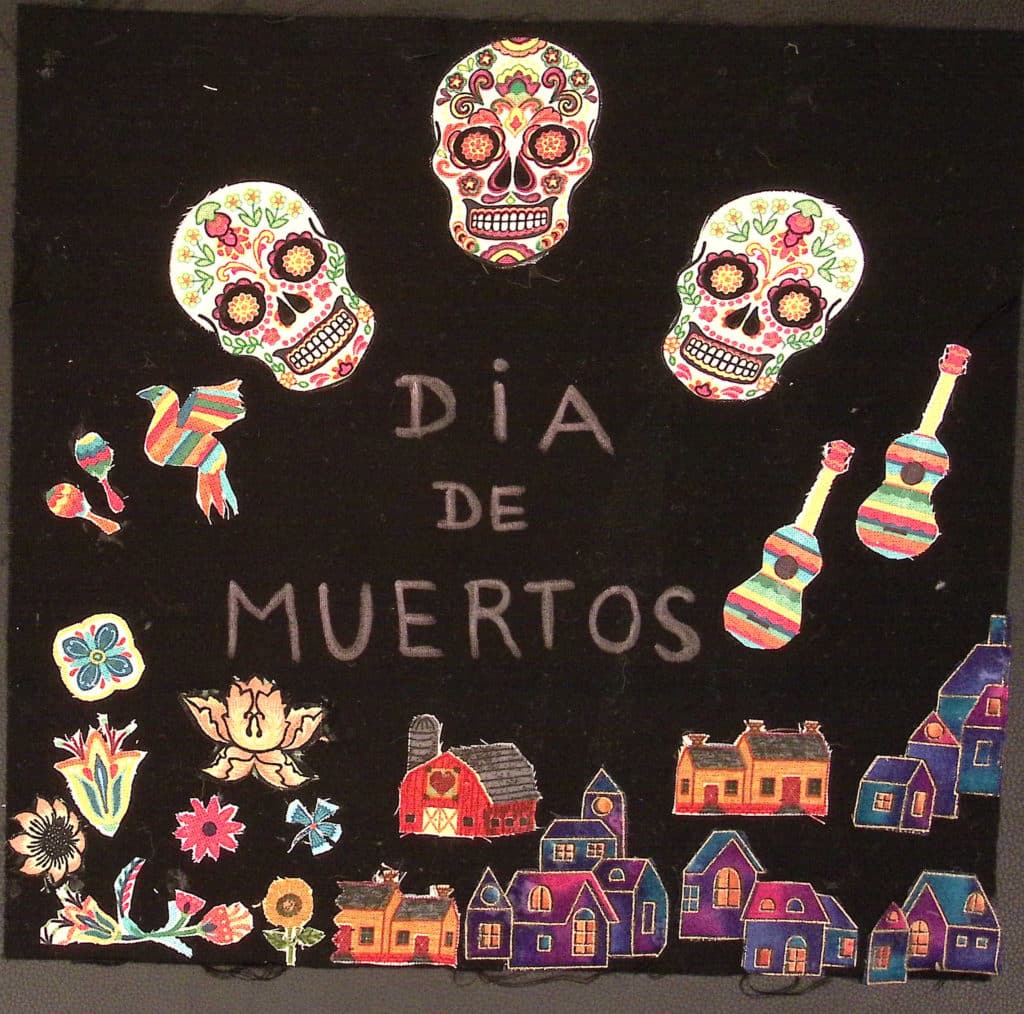 Black square with three decorated skulls and the words "Dia de Muertos," with houses and guitars.