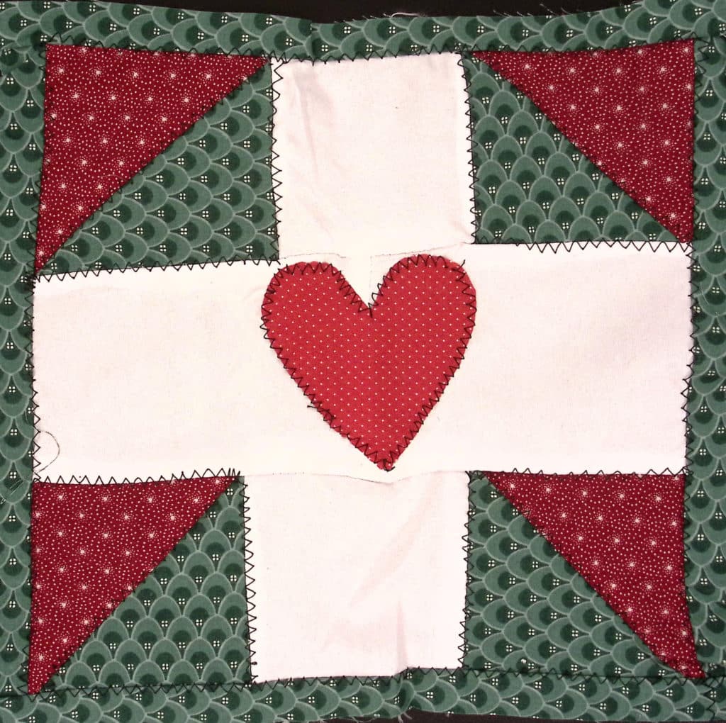 A quilted green and red square with a white cross and a heart in the center.
