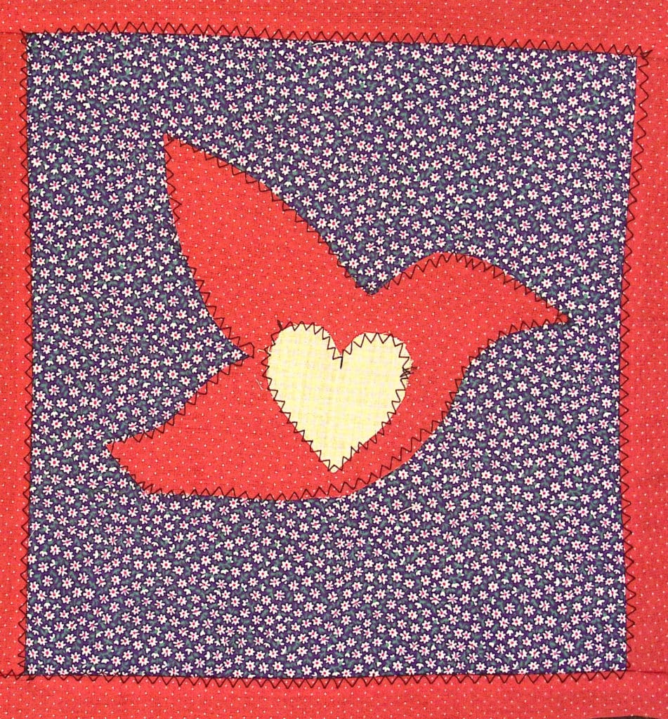 A quilted square featuring a red bird with a white heart in the middle.