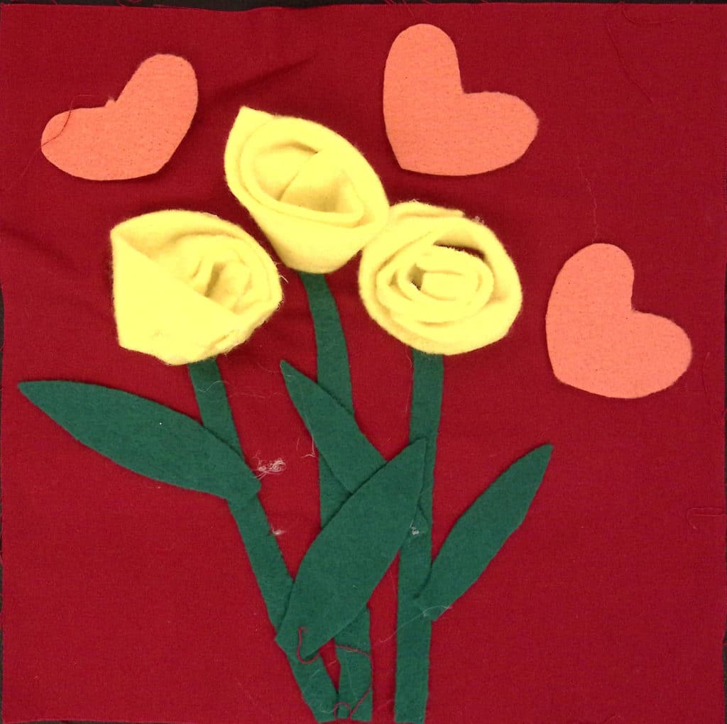 Yellow felt roses with green stems, surrounded by pink hearts on a red background