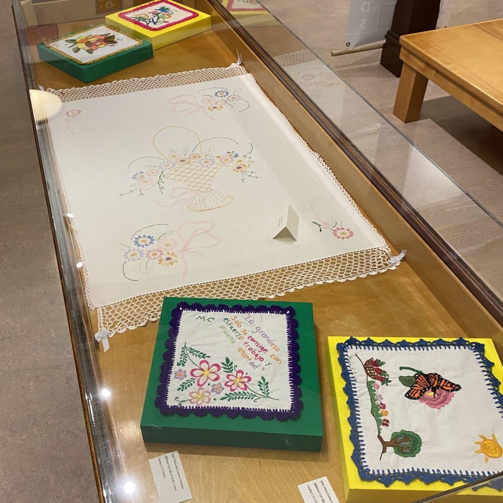 Exhibit case showing four embroidered quilt squares and a table runner embroidered with a basket of flowers