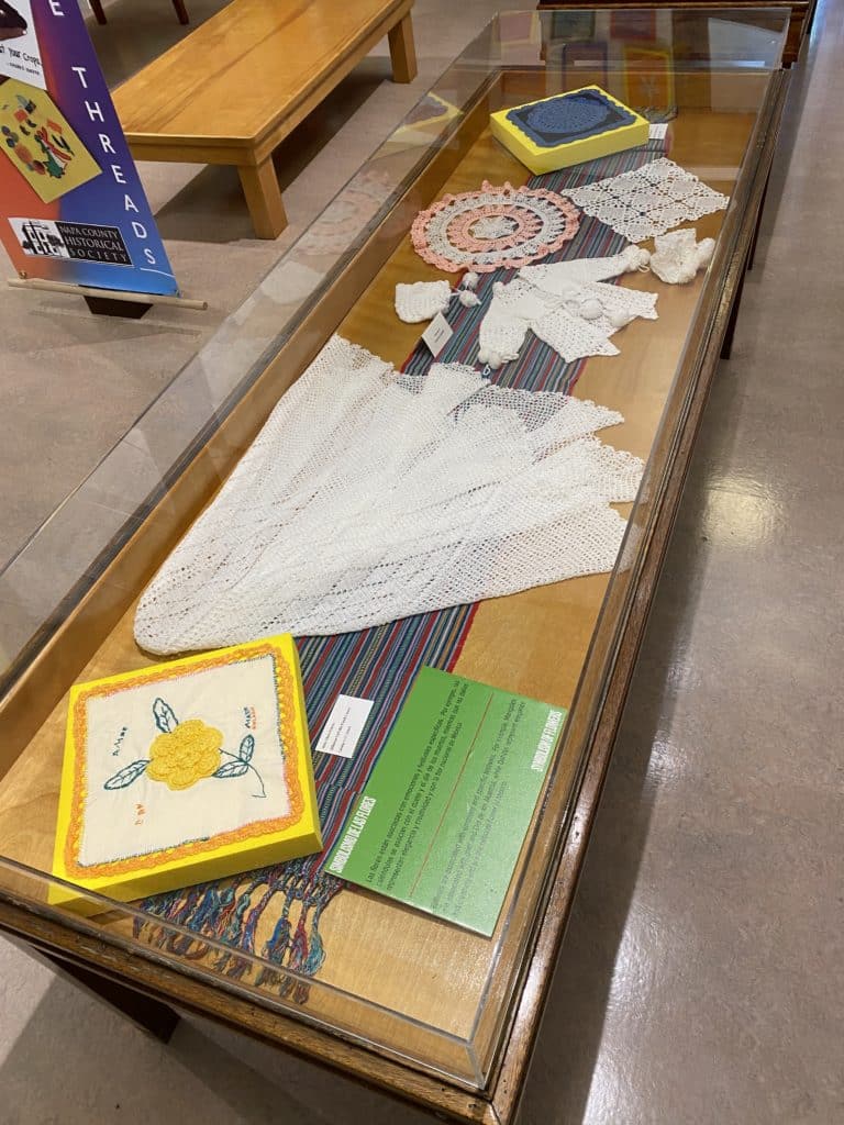Exhibit case displaying crocheted baby clothes, doilies, and two quilt squares featuring crocheted pieces.