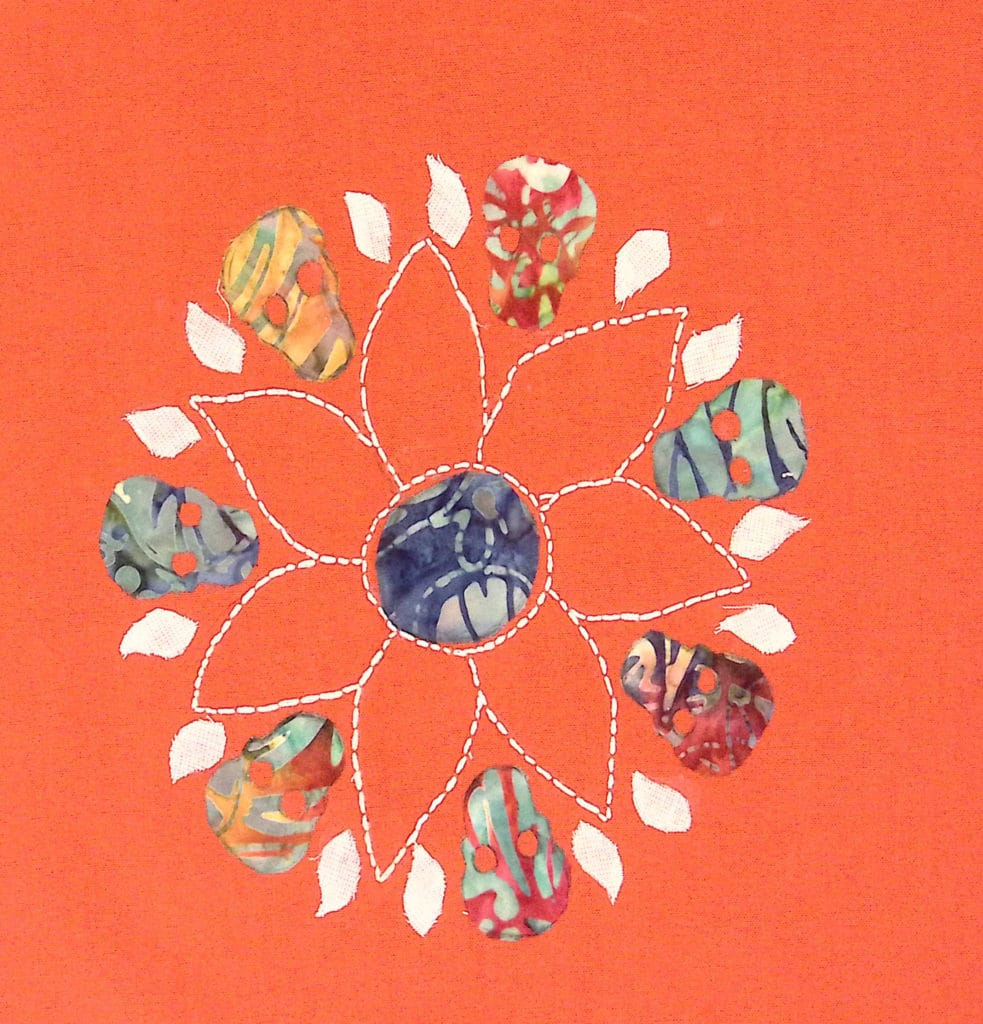 An orange background with white embroidered flower and tie-dye pattern skulls arranged in a mandala shape