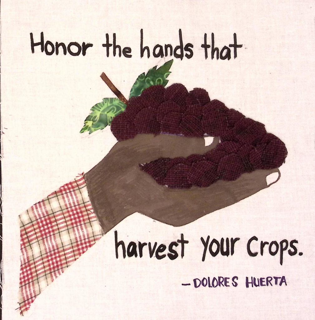 A hand, with a red plaid sleeve, holds out dark purple grapes. Text surrounding the image reads, "Honor the hands that harvest your crops. -Dolores Huereta."