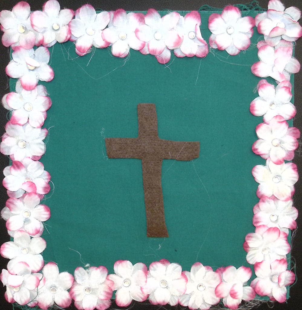 A brown cross on a green background, with pink and white silk flowers around the edge