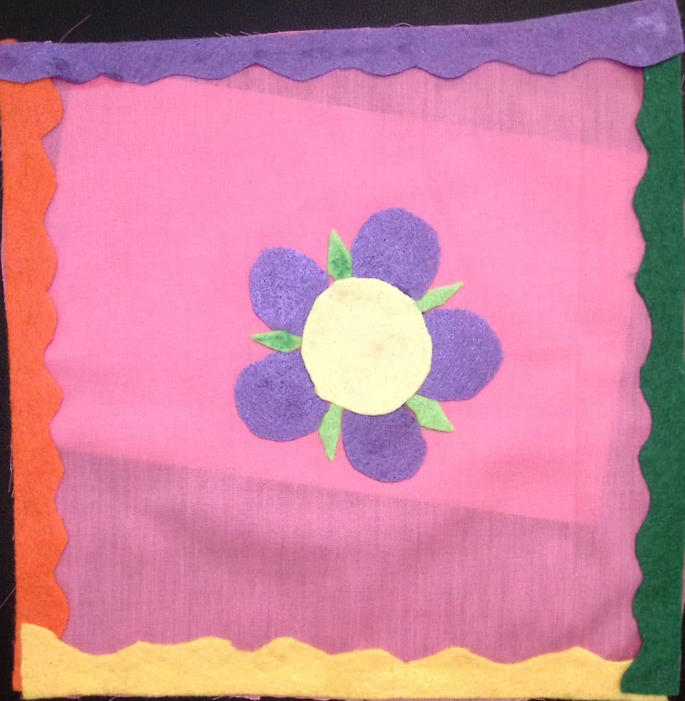 A purple and green felt flower on a pink background with scalloped multicolor borders
