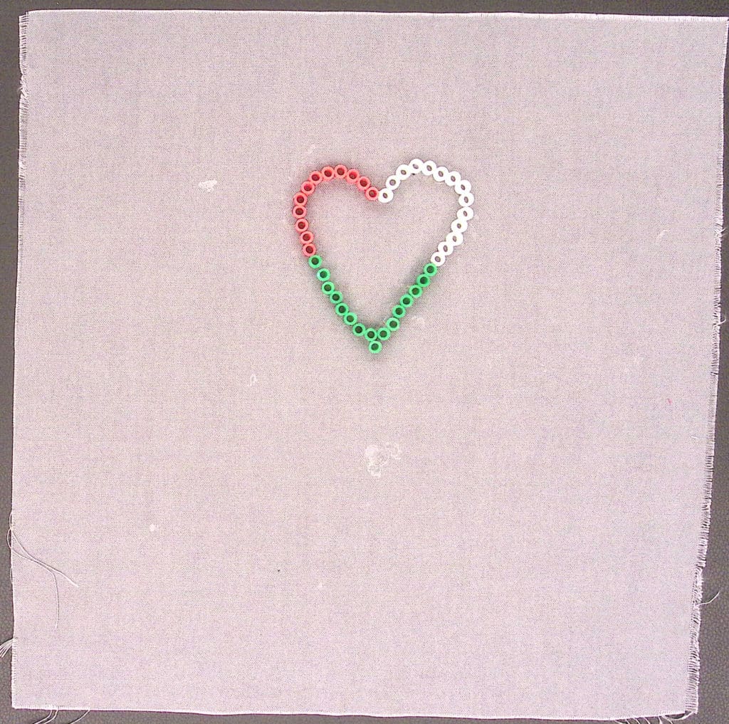 A green, red, and white heart made with perler beads on a gray square.