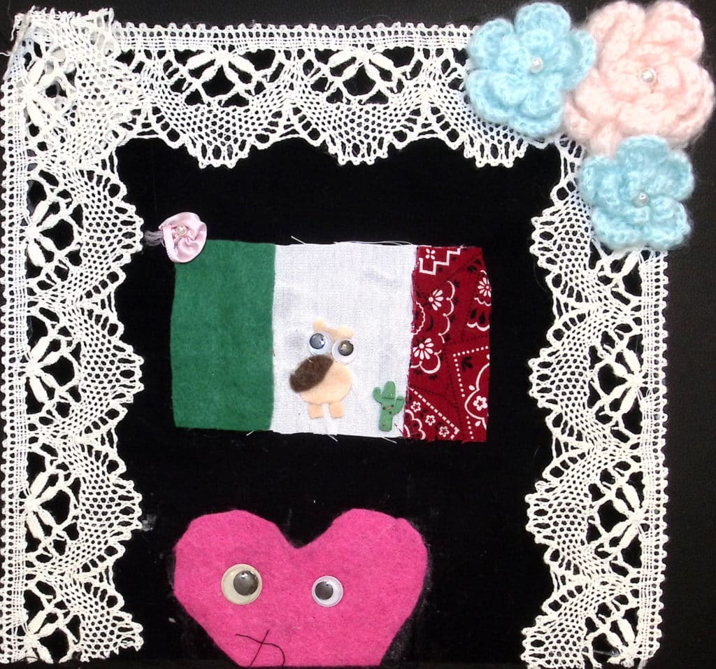 A Mexican flag and a pink heart with googly eyes, with white lace border and crocheted blue and pink flowers in corner.