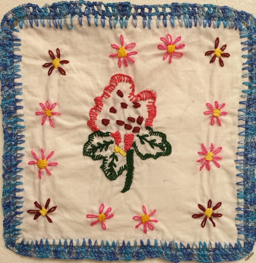Embroidered pink flower with green leaves, smaller pink and purple flowers along border.