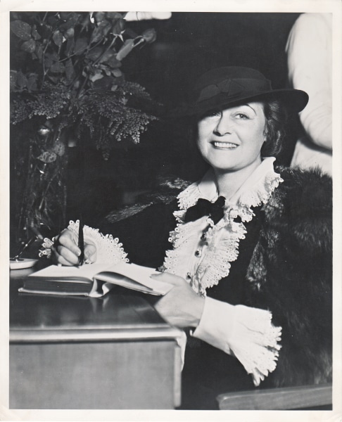 Frances Marion sitting at a desk with a pen in her hand, possibly signing a book.