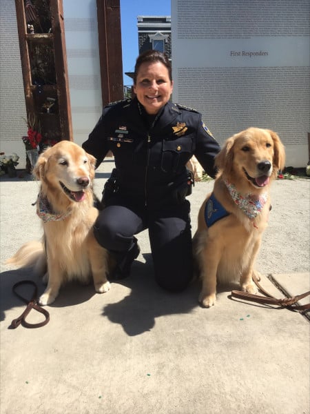 Jennifer Gonzales in her police uniform, seated with two golden retriever dogs in front of the Napa 9/11 Memorial.