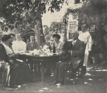 Eight people seated around a table on a patio, friends or family of the Lymans. Ah Hing, the cook and manager of the Lyman household, stands behind them wearing a white coat or apron.