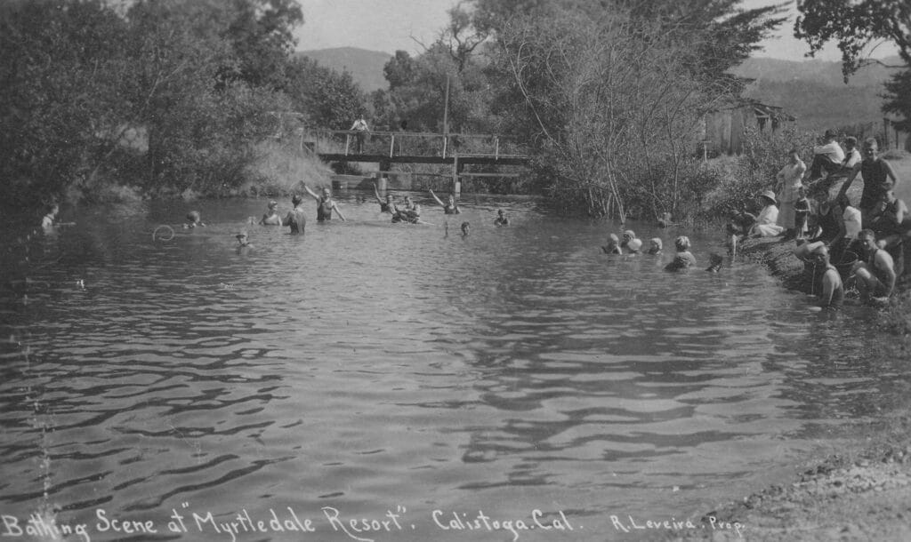 People swimming in the river at Myrtledale Resort. One man stands on a bridge above the river.