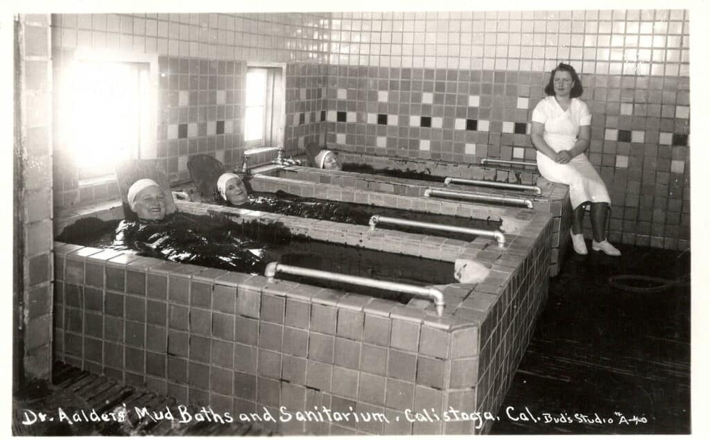 Women in the mudbaths at Dr. Aalder's, one woman dressed as an attendant sits on the side of the tubs.